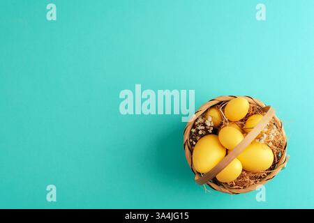 Korb mit gelben Ostereiern, Stroh und getrockneten Blumen auf pastellfarbenem türkisfarbenem Hintergrund. Draufsicht, flach, Kopierbereich Stockfoto