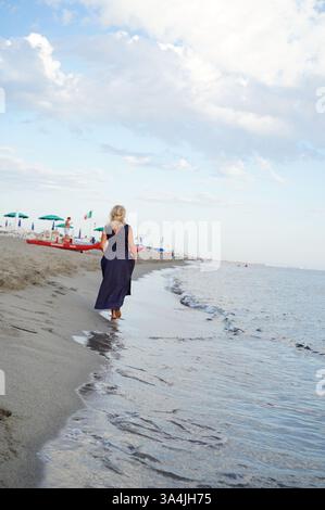 Eine Person in dunklem Kleid spaziert entlang eines Sandstrandes und hinterlässt Spuren im nassen Sand, während sanfte Wellen über die Küste ziehen. Der Himmel ist teilweise bewölkt, Indicati Stockfoto