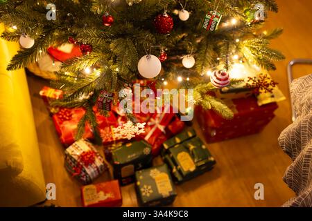 Ein wunderschön dekorierter Weihnachtsbaum mit leuchtenden Lichtern und festlichen Ornamenten steht über einer Sammlung bunter Geschenkverpackungen. Stockfoto