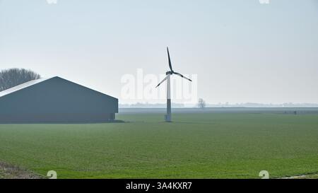 10. März 2025 - Groningen-Niederlande: In der Nähe eines traditionellen niederländischen Bauernhofs steht eine kleine Windkraftanlage, die erneuerbare Energie in einer ländlichen Landschaft erzeugt. Stockfoto