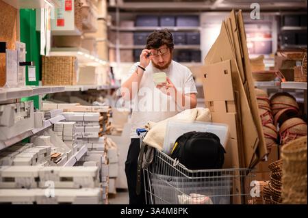 Ein Mann in einem weißen T-Shirt untersucht ein Produkt beim Einkaufen von Essentials. Er steht in der Nähe von Regalen, die mit Haushaltsgegenständen bestückt sind, mit vollem Wagen Stockfoto