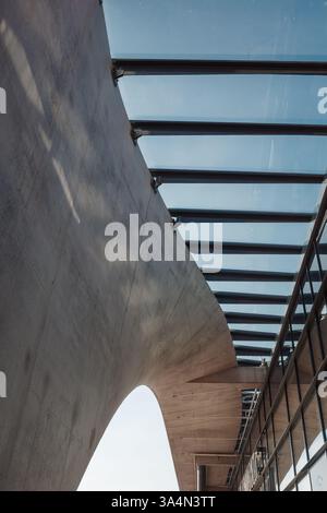 Eine moderne architektonische Struktur mit einem eleganten Glasdach, das von Metallbalken gestützt wird und von geschwungenen Beton- und Ziegelwänden eingerahmt wird. Stockfoto