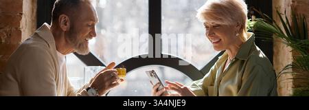 Ein Paar mittleren Alters erzählt Lachen und Geschichten bei Getränken, während es auf ein Smartphone und Banner blickt Stockfoto