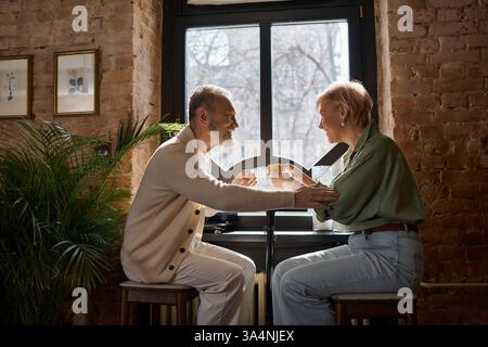 Ein Paar mittleren Alters lacht und trinkt und genießt ihre intime Zeit am Fenster. Stockfoto