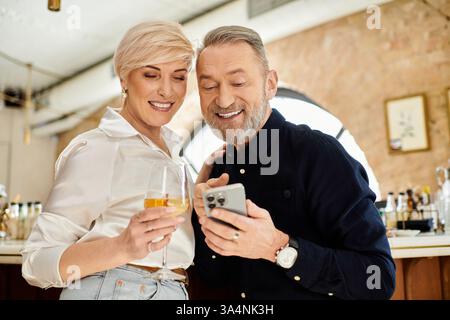 Ein Paar mittleren Alters lacht, während es auf ein Telefon schaut und ihr Date mit Drinks genießt. Stockfoto