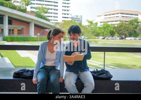 Zwei asiatische Studenten saßen auf einer Bank in einem modernen Universitätsumfeld und nahmen an einer freundlichen Diskussion über ein gemeinsames Buch Teil Stockfoto