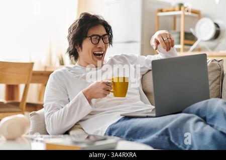 Ein fröhlicher Student lernt auf seinem Laptop und schlürft in einer komfortablen Wohnumgebung ein warmes Getränk. Stockfoto