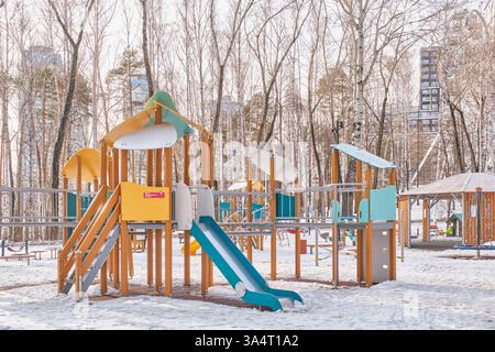 Jekaterinburg, Russland - 17. März 2025: Moderner Spielplatz mit Schnee. Bunte Rutschen und Holzelemente. Umgeben von blattlosen Bäumen mit hohen Bäumen Stockfoto