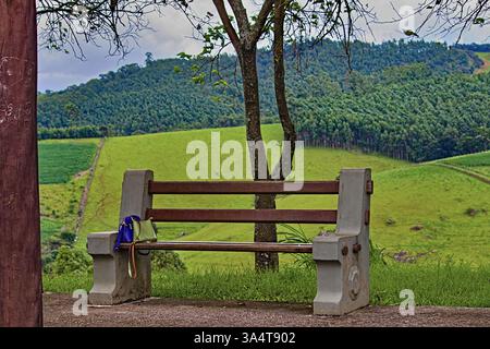 Bank mit Holzsitz und Zementstruktur, mit zwei Damenhandtaschen links auf der Seite und im Hintergrund Hügel mit grünen Feldern und Strecken von Stockfoto