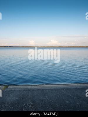 Sonnenuntergang über dem Meer: Wenige Wolken spiegeln sich im Wasser über der leeren Bucht mit Betondamm. Stockfoto