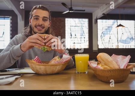 Junger Mann, der Burger und Orangensaft in einem gemütlichen Café genießt, glücklich lächelnd, Kopierraum Stockfoto