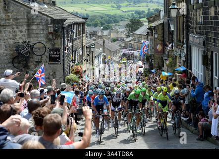 Dateifoto vom 07/14 vom Hauptfeld fährt die Main Street hinauf, während die zweite Etappe der Tour de France durch Haworth, Yorkshire, führt. Edinburgh wurde ausgewählt, um eine „herrliche Kulisse“ für den Beginn der Tour de France 2027 zu bieten. Schottland, England und Wales teilen sich sechs Etappen des weltweit größten Radsports – drei für Männer- und Frauenrennen, die beide zum ersten Mal im gleichen Land, dem Vereinigten Königreich, starten werden. Ausgabedatum: Mittwoch, 19. März 2025. Stockfoto