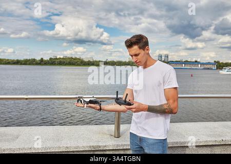 Ein junger Mann mit einer Drohne und ihrem Controller steht auf einem Damm am Fluss unter einem teilweise bewölkten Himmel. Stockfoto