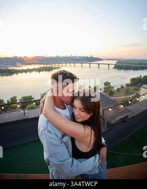 Ein junges Paar genießt einen ruhigen Abend auf einem Dach, umgeben von den warmen Farbtönen der untergehenden Sonne. Ihre Umarmung spricht von gemeinsamen Momenten und Ruhe Stockfoto