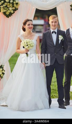 Elegante Hochzeitsszene, die die Braut mit einem weißen Rosenstrauß, den Bräutigam in einem klassischen Anzug und einen Trauzeugen zeigt, der auf einem weißen Teppich unter einem steht Stockfoto