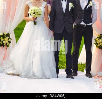 Elegante Hochzeitsszene, die die Braut mit einem weißen Rosenstrauß, den Bräutigam in einem klassischen Anzug und einen Trauzeugen zeigt, der auf einem weißen Teppich unter einem steht Stockfoto