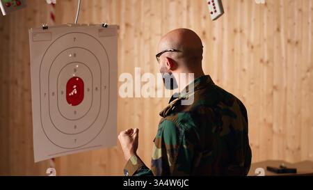 Armeesoldat im Schießstand lädt Pistole, Schießziel, Testgenauigkeit, Ergebnisse. Militärmann auf Schießstand übt mit Waffe und überprüft, ob er Bullseye erschossen hat, Kamera B Stockfoto