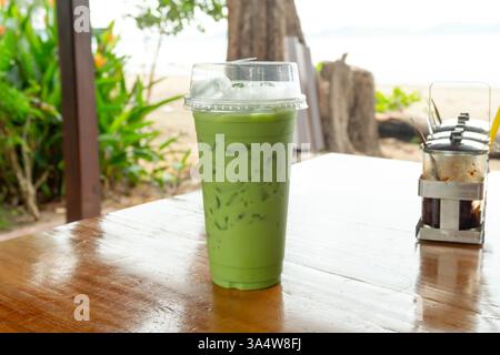 Ein kaltes Getränk aus eiskaltem Matcha Latte Grüntee mit Kondensmilch in transparentem Plastiktrinkglas ist auf dem Tisch des draußen gelegenen Cafés in der Nähe des Meeres. Stockfoto