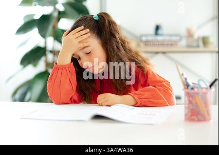 Müdigkeit ein weißes Schulmädchen in roter Bluse sitzt mit geschlossenen Augen an einem weißen Schreibtisch, legt ihre Stirn auf ihre Hand, langweilig und frustriert während der Hausaufgaben zu Hause Stockfoto