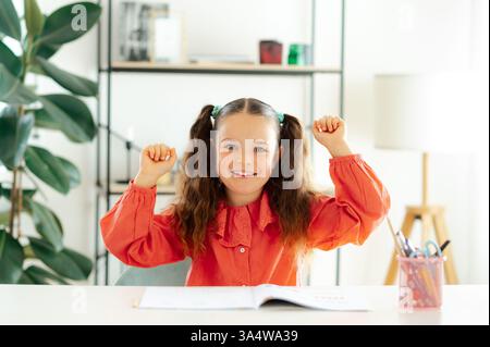 Glückliches süßes weißes blauäugiges Mädchen 6-7 Jahre mit langen lockigen Haaren in einer roten Bluse, das an einem Schreibtisch sitzt, ihre Hände hochhebt und sich glücklich in die Kamera lächelt. Hausaufgaben zu Hause machen Stockfoto