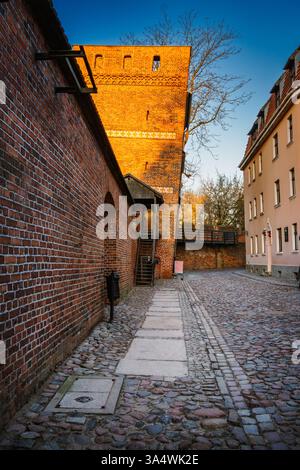 Während die Sonne untergeht, strahlt warmes Licht über dem Schiefen Turm in Torun und beleuchtet die kopfsteingepflasterte Straße und mittelalterliche Architektur. Stockfoto