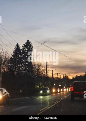 Überqueren Sie den Himmel der Wolken oder Chemtrails in der Abenddämmerung an einem düsteren, kühlen Abend über Autos mit Scheinwerfern, die auf einer von Bäumen gesäumten Straße fahren. Stockfoto