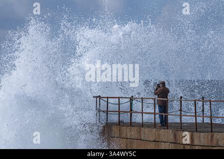 Ein Beobachter auf dem Pier in Porthleven wird fast nass, als eine Welle vor ihm bricht und überall Sprühnebel auftaucht. Stockfoto