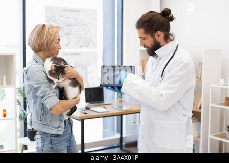 Tierarzt erklärt Röntgenbild einer jungen Frau, die ihre Hauskatze im Büro hält. Bild geeignet für Gesundheit, tierärztliche Dienste und Tierschutz. Stockfoto
