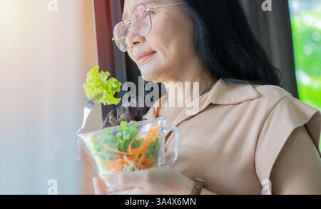 Nahaufnahme einer lächelnden erwachsenen asiatischen Frau, die im Wohnzimmer einen Salat isst. Schöne gesunde Frau, die Salat isst. Kopierbereich. Stockfoto