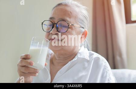 Glückliche lächelnde asiatische Seniorin trinkt warme Milch oder Nahrungsergänzungsmittel aus dem Glas. Selektiver Fokus. Stockfoto