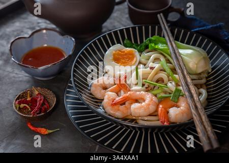 Udon-Nudeln mit Ei, Garnelen, Pilzen, roter Chili, auf dunklem rustikalem Hintergrund mit Teetassen und Sauce Stockfoto
