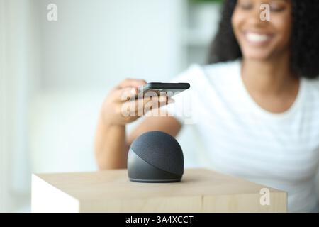Glückliche schwarze Frau, die ihr Telefon mit intelligentem Lautsprecher verbindet und zu Hause auf einer Couch sitzt Stockfoto