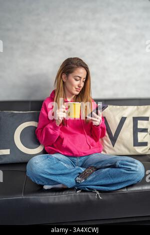 Junge Frau, die bequem auf einem Sofa sitzt, eine Tasse Tee genießt und ihr Smartphone durchsucht Stockfoto