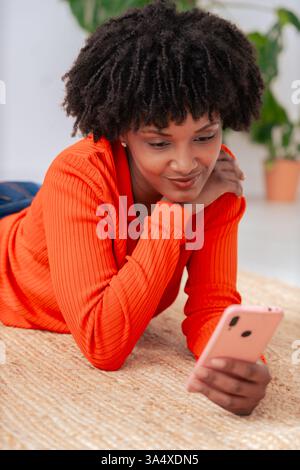 Glückliche Afroamerikanerin, die ihr Smartphone benutzt, während sie zu Hause auf dem Boden liegt. Stockfoto