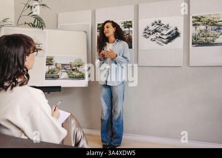 Eine Frau im mittleren Alter leitet eine Projektmanagementsitzung, in der sie über ausgestellte Architekturentwürfe diskutiert. Sie engagiert ihren Kollegen mit visuellen Plänen für eine M Stockfoto