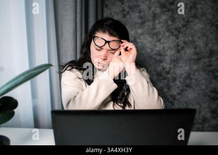 Symptome der Überlastung. Junge müde Freiberuflerin in Brille, die vor Computer sitzt und die Augen während der Arbeit anstrengt. Stockfoto
