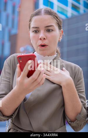 Schockierte kaukasische Geschäftsfrau, die draußen mit überraschtem Ausdruck auf das Smartphone blickt. Stockfoto