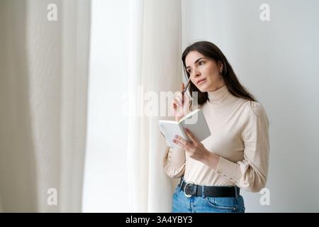 Die Frau schaut sich ein Buch an und schreibt darin. Sie trägt ein weißes Hemd und blaue Jeans Stockfoto