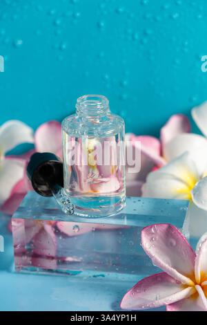 Ein klares Serum in einer Tropfflasche liegt auf einem Glasblock auf, der mit zarten Plumerieblättern verziert ist. Stockfoto