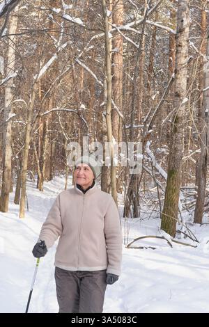 Seniorin Skilanglauf durch verschneite Wälder. Sie lächelt, umgeben von schneebedeckten Bäumen. Sonnenlicht strömt durch die Äste. Ruhig o Stockfoto