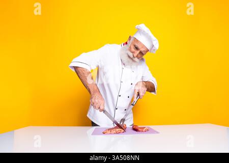 Der erfahrene Koch bereitet Gourmetmahlzeiten auf leuchtendem gelbem Hintergrund in traditioneller Uniform und zielgerichtetem Ausdruck zu Stockfoto