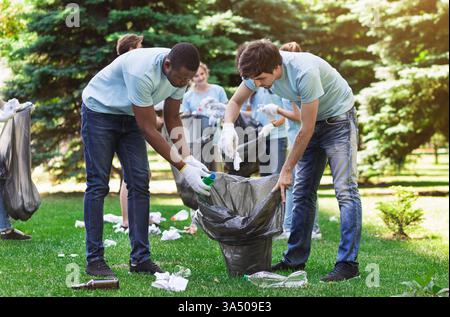 Verschiedene Freiwillige in blauen Hemden und Handschuhen sammeln recycelbare Plastikflaschen in einem Park. In dieser Szene werden Umweltschutz, Teamarbeit und Freiwilligenarbeit im Freien hervorgehoben. Stockfoto