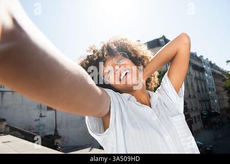 Fröhliches Porträt einer jungen Afroamerikanerin, die ein Selfie in einer städtischen Umgebung macht. Sie verströmt Selbstvertrauen und einen lebendigen Lebensstil in der Stadt, mit einem Outdoor-Stadtbild hinter sich. Ideal für Lifestyle, urbane Mode und Straßenfotografie. Stockfoto