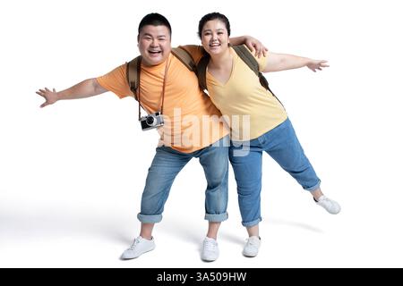 Fröhliches chinesisches Paar in Übergröße mit Rucksäcken steht mit Armen umeinander und ausgestreckten Armen in einem Studio vor weißem Hintergrund. Ein vielseitiges Image für Reisen, Paare und Lifestyle-Kampagnen, das Glück und Zweisamkeit hervorhebt. Stockfoto