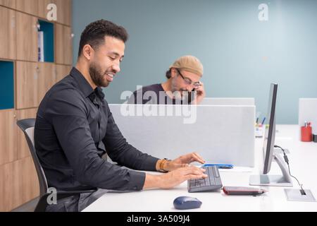 Lächelnder Geschäftsmann aus dem Nahen Osten, der auf der Tastatur sitzt, während er am Tisch neben einem männlichen Kollegen sitzt und im Büro telefoniert Stockfoto