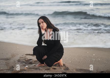 Gemischte Rassenfrau mit langen dunklen Haaren, die ein schwarzes Maxi-Kleid trägt und wegschaut, während sie sich während des Urlaubs mit Hand auf Wange am Sandstrand hockt Stockfoto