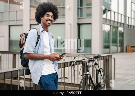 Ein lächelnder Afroamerikaner mit Afro und Schnurrbart steht neben einem Fahrrad vor einem Metallzaun in einer belebten Stadtstraße. Dieses ansprechbare Stadtporträt eignet sich für Lifestyle-, Pendler- und Straßenfotografie. Stockfoto