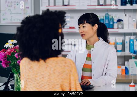 Porträt einer vielfältigen Apothekenszene mit einer asiatischen Apothekerin und einer afroamerikanischen Kundin, die Medikamente am Tresen diskutieren. Das Bild vermittelt professionelle Betreuung und Patientenberatung in einem Drogeriemarkt. Kopierbereich verfügbar für Overlays und Nachrichten. Stockfoto