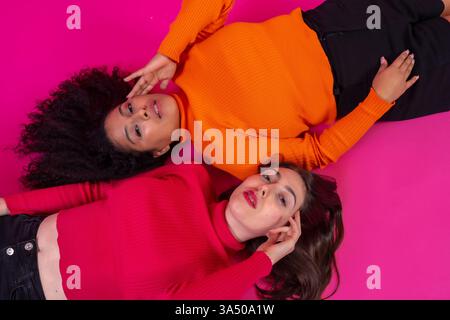 Zwei Frauen gemischter Herkunft liegen nahe auf einem rosa Studioboden und berühren ihre Gesichter in spielerischer Pose. Eine unterstützende, vielfältige Freundschaftsszene, die zu Beauty-, Lifestyle- und Wellness-Kampagnen passt. Ideal für Inclusive Marketing- und Hautpflegebotschaften. Stockfoto