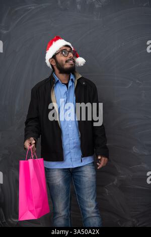 Indischer Mann in festlicher Partykleidung, trägt einen Weihnachtsmann-Hut und hält eine Einkaufstasche vor einem dunklen Studiohintergrund. Dieses Weihnachtsbild fängt Weihnachtsfreude und saisonale Geschenke für Weihnachtskampagnen ein. Ideal für Einzelhändler, die festliche Verkäufe während der Feiertage fördern. Stockfoto